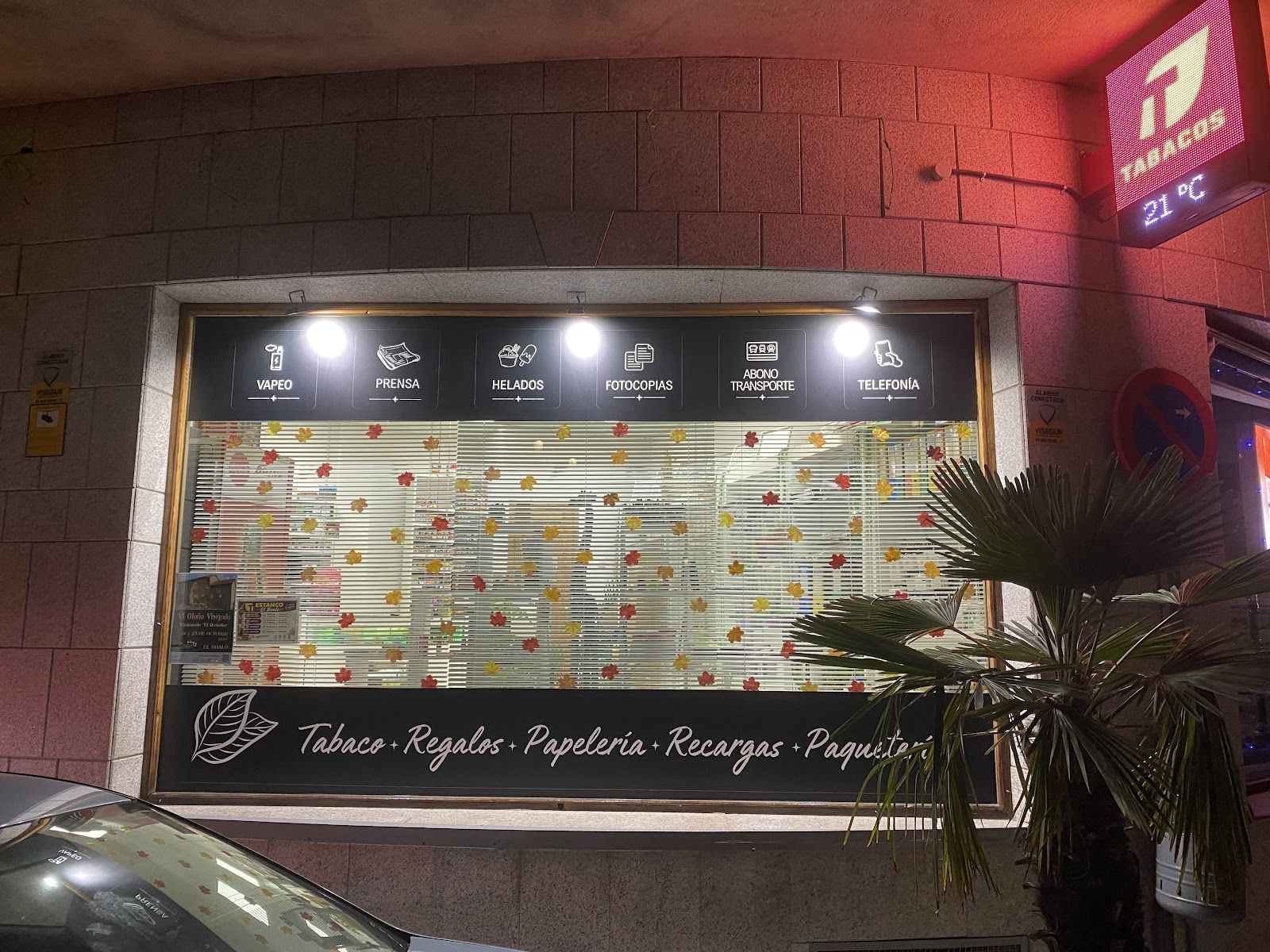 Escaparate iluminado de noche con los servicios: vapeo, prensa, helados, fotocopias, abono transporte y telefonía